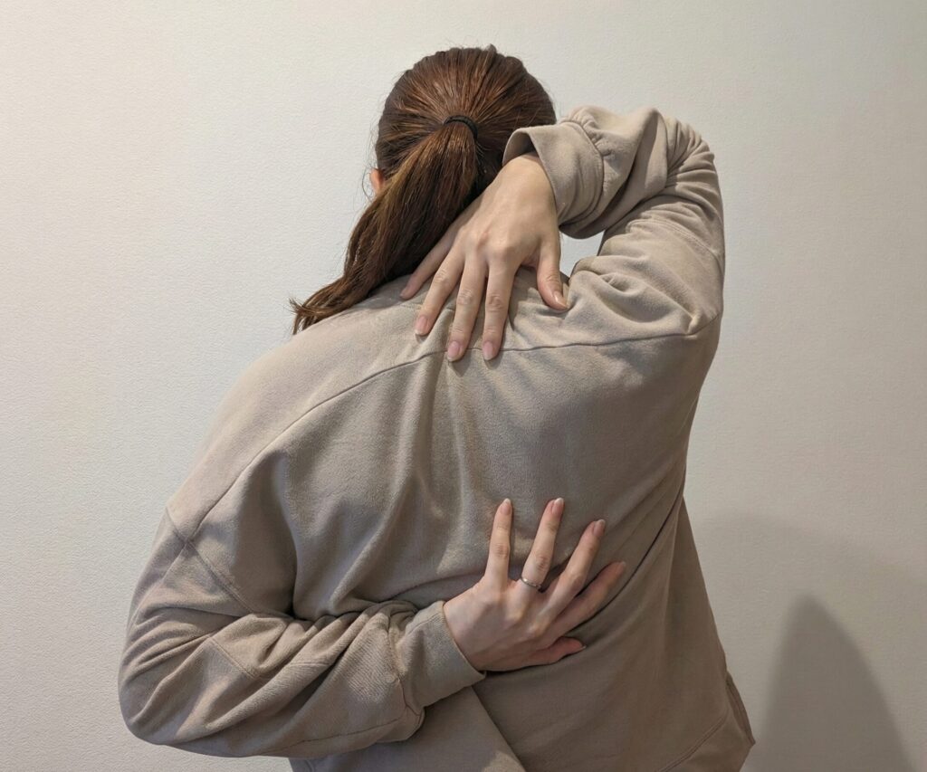 ベージュのスウェットシャツを着て、髪をポニーテールに結んだ女性の後ろ姿。背中側で左手を上から、右手を下から回し、両手の指を組もうとするストレッチのポーズをとっている。両手の指先はまだ届いておらず、現時点で指と指の間の距離は13cm空いている状態を示す写真。